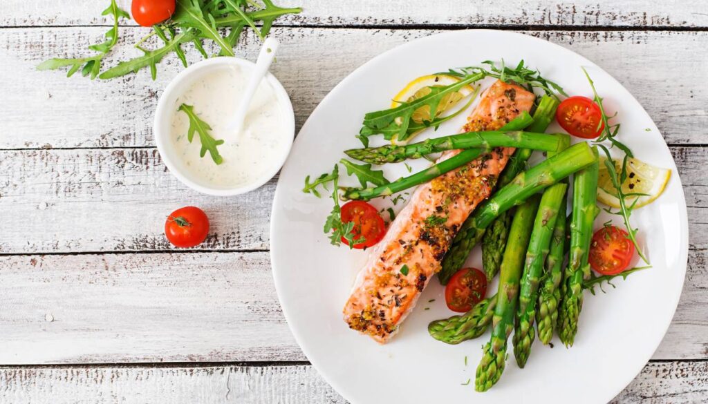 A plate of herb-crusted salmon filet served with asparagus, cherry tomatoes, and lemon slices, garnished with arugula, accompanied by a small bowl of creamy sauce.