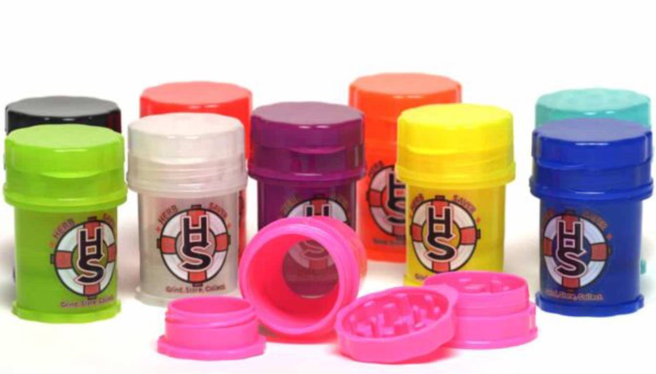 A collection of colorful Herb Saver grinders in various colors including green, white, purple, yellow, blue, and pink. One pink grinder is open, showing its internal grinding mechanism.