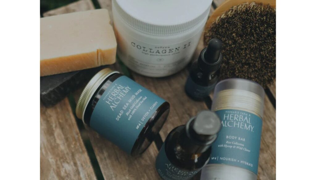 A collection of natural skincare products from Herbal Alchemy, including a Dead Sea Mud Mask, a Body Bar, a Collagen II jar, and a soap bar, arranged on a wooden surface with a brush.