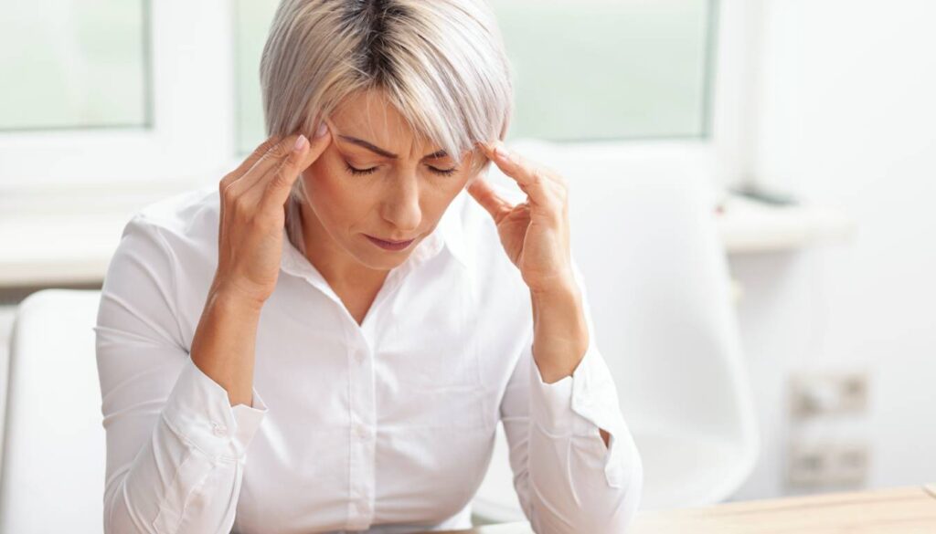 A person in a white shirt sitting at a desk, holding their head with both hands, possibly experiencing a headache or migraine.