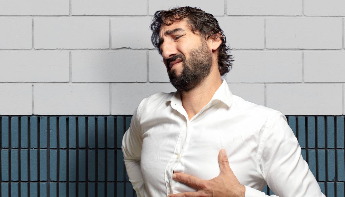 A person in a white shirt holding their chest, standing against a background of white and blue brick walls.