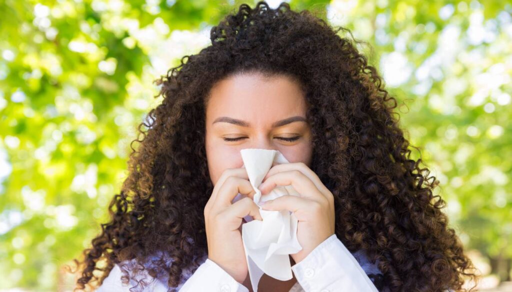 Person with curly hair holding a tissue to their nose, standing outdoors with a blurred green background.