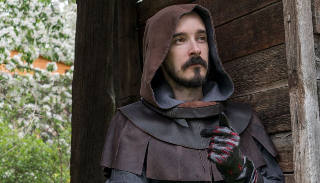 A person wearing a brown hooded cloak and black gloves stands in front of a wooden structure, with greenery and white flowers in the background.