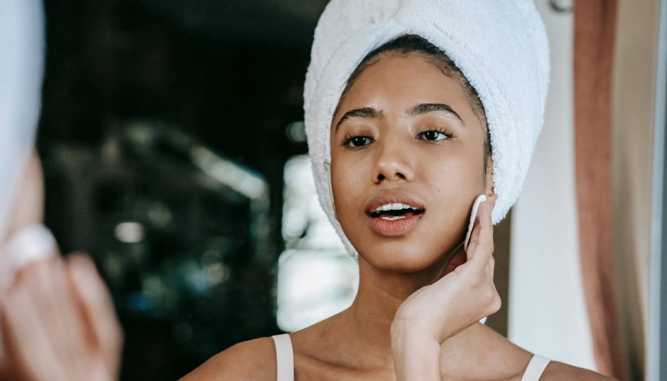 Person with a towel wrapped around their head, applying skincare in front of a mirror.