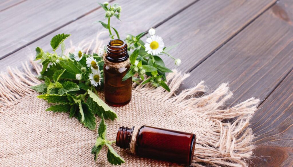 A close-up of two small brown glass bottles, one standing and one lying down, surrounded by fresh herbs and small white flowers, placed on a rustic burlap cloth on a wooden surface.