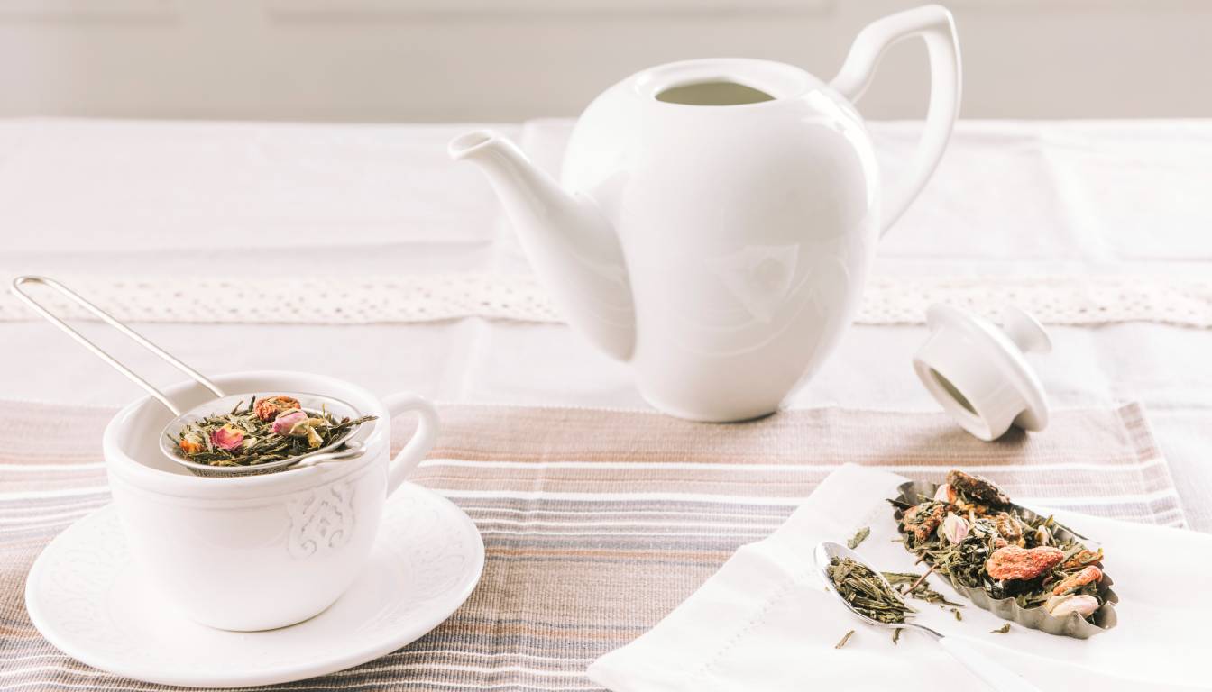 A white teapot with its lid off sits on a table next to a white teacup with a metal tea infuser filled with herbal tea leaves and flowers. A spoon and additional herbal tea leaves are placed on a white napkin nearby.