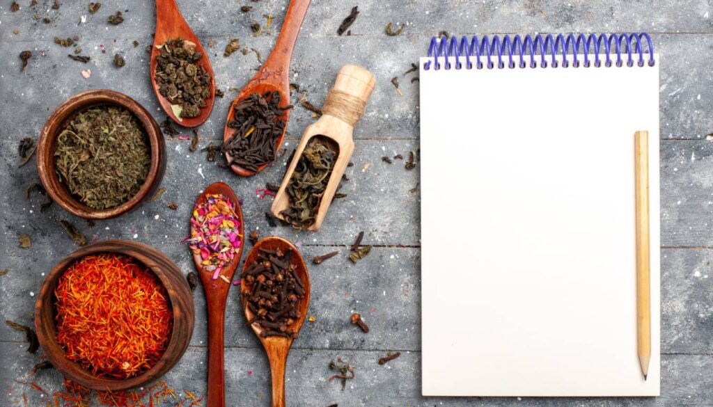 Various dried herbs and spices in wooden bowls and spoons arranged on a rustic surface next to a blank notepad with a pencil.