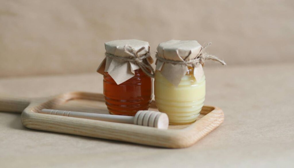 Two jars of honey, one dark and one light, with paper covers and twine ties, placed on a wooden tray with a honey dipper.