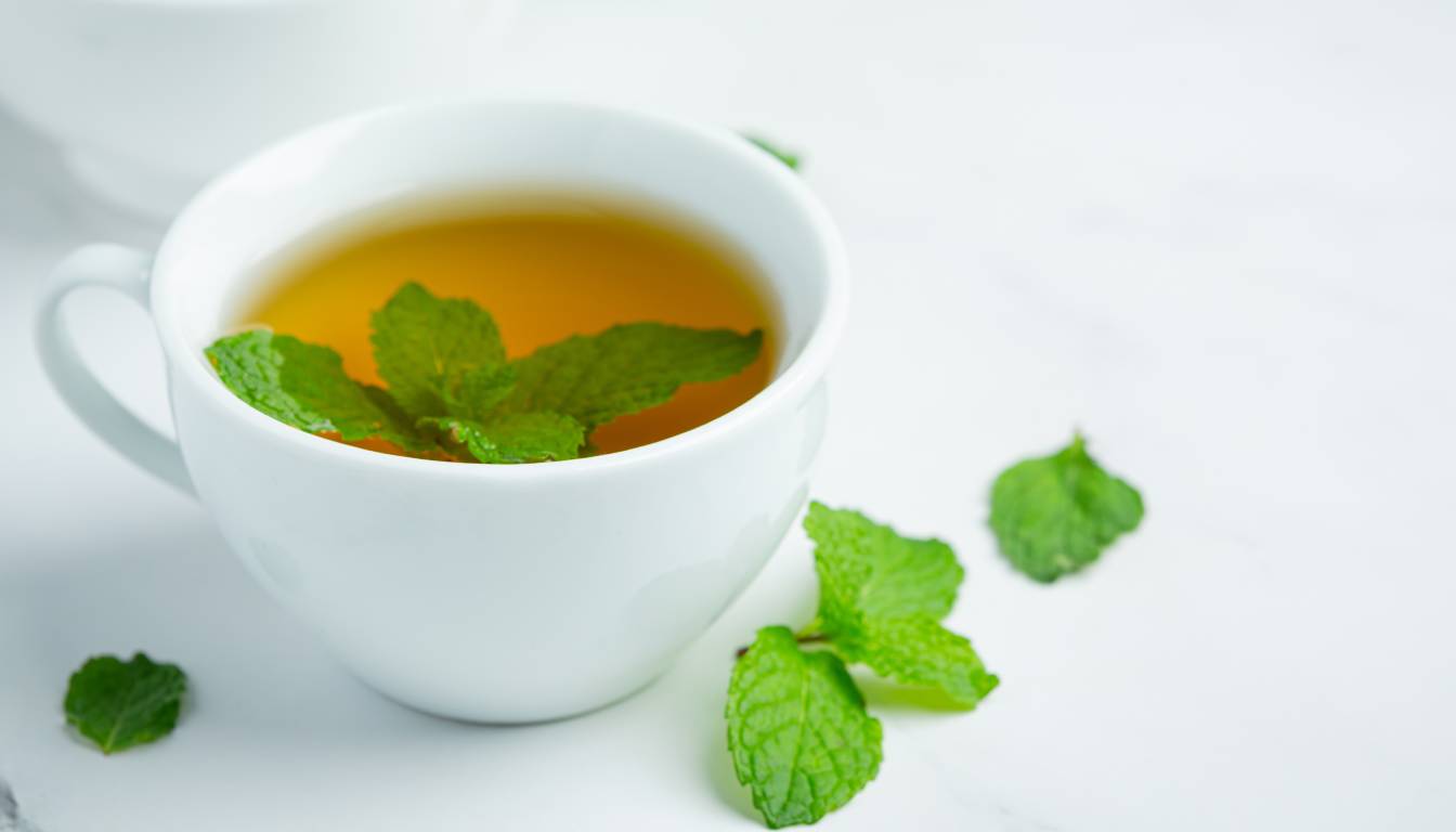 A white cup filled with peppermint tea, garnished with fresh mint leaves, placed on a white surface with additional mint leaves scattered around.