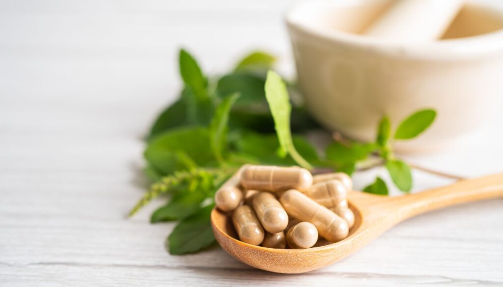 A wooden spoon filled with herbal supplement capsules, surrounded by fresh green leaves and a mortar and pestle in the background.