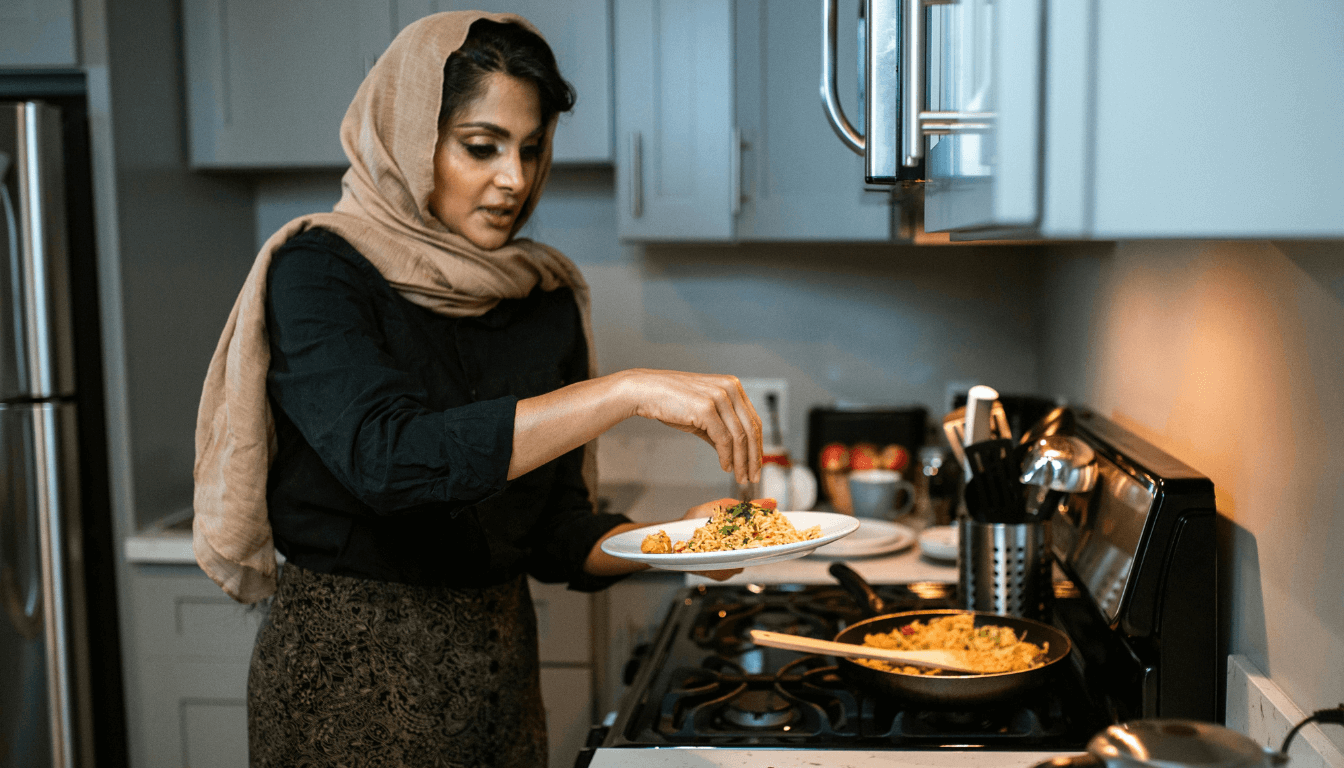 An individual in a kitchen, facing away from the camera, garnishing a dish with herbs. The individual is wearing a headscarf and standing in front of a stove with a pan of food.