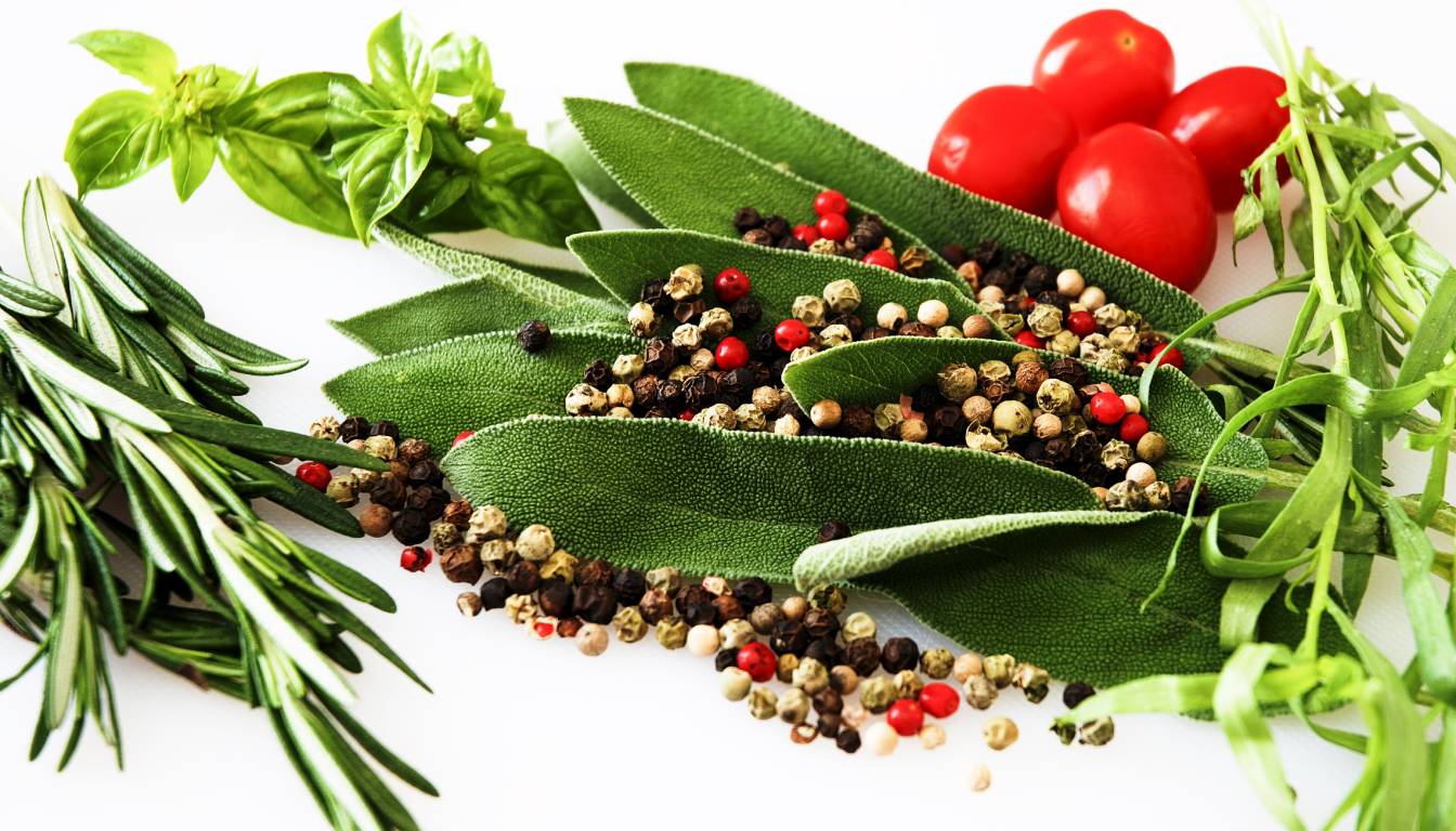 A variety of fresh herbs and spices including basil, rosemary, sage, and tarragon, along with whole peppercorns and cherry tomatoes, arranged on a white background.