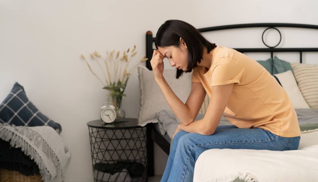 A person sitting on a bed with a hand on their forehead, appearing to be in deep thought or distress. The room is decorated with pillows, a bedside table with a clock, and a vase with dried flowers.