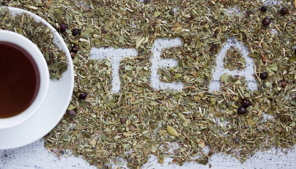 A white cup filled with herbal tea sits on a saucer, surrounded by loose herbal tea leaves. The word "TEA" is spelled out beneath the leaves.