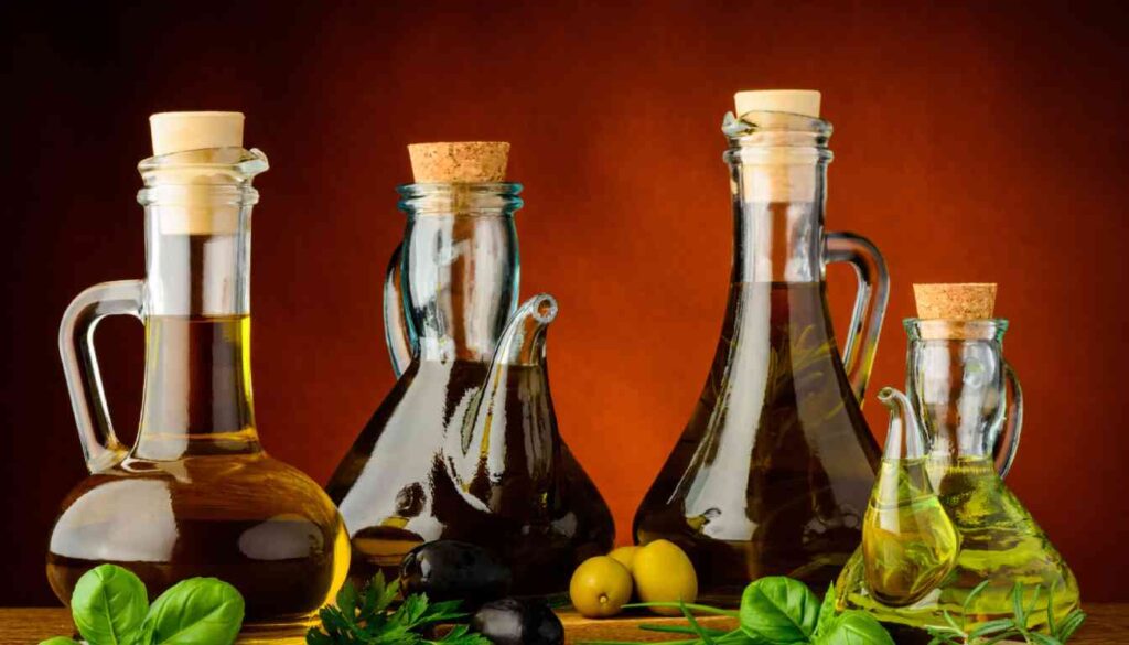 Four glass bottles of herb-infused olive oil with cork stoppers, surrounded by fresh herbs and olives, against a warm, reddish-brown background.