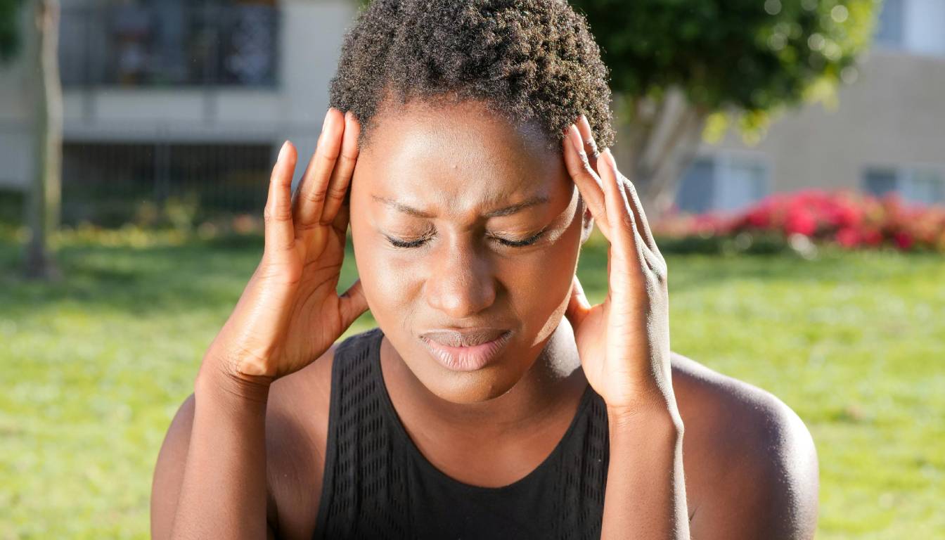 A person holding their head with both hands, possibly indicating a headache or migraine, while sitting outdoors on a sunny day.