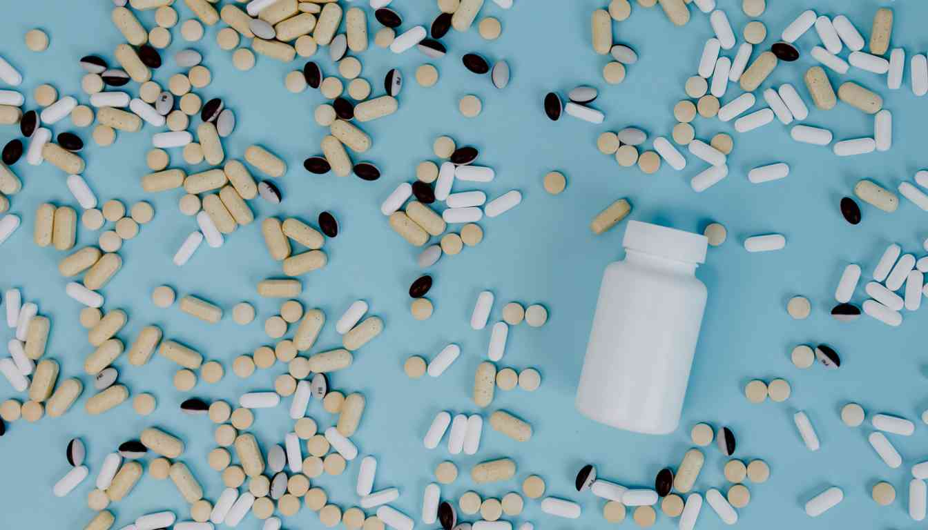 A variety of pills and capsules scattered on a light blue surface with a white bottle lying on its side.