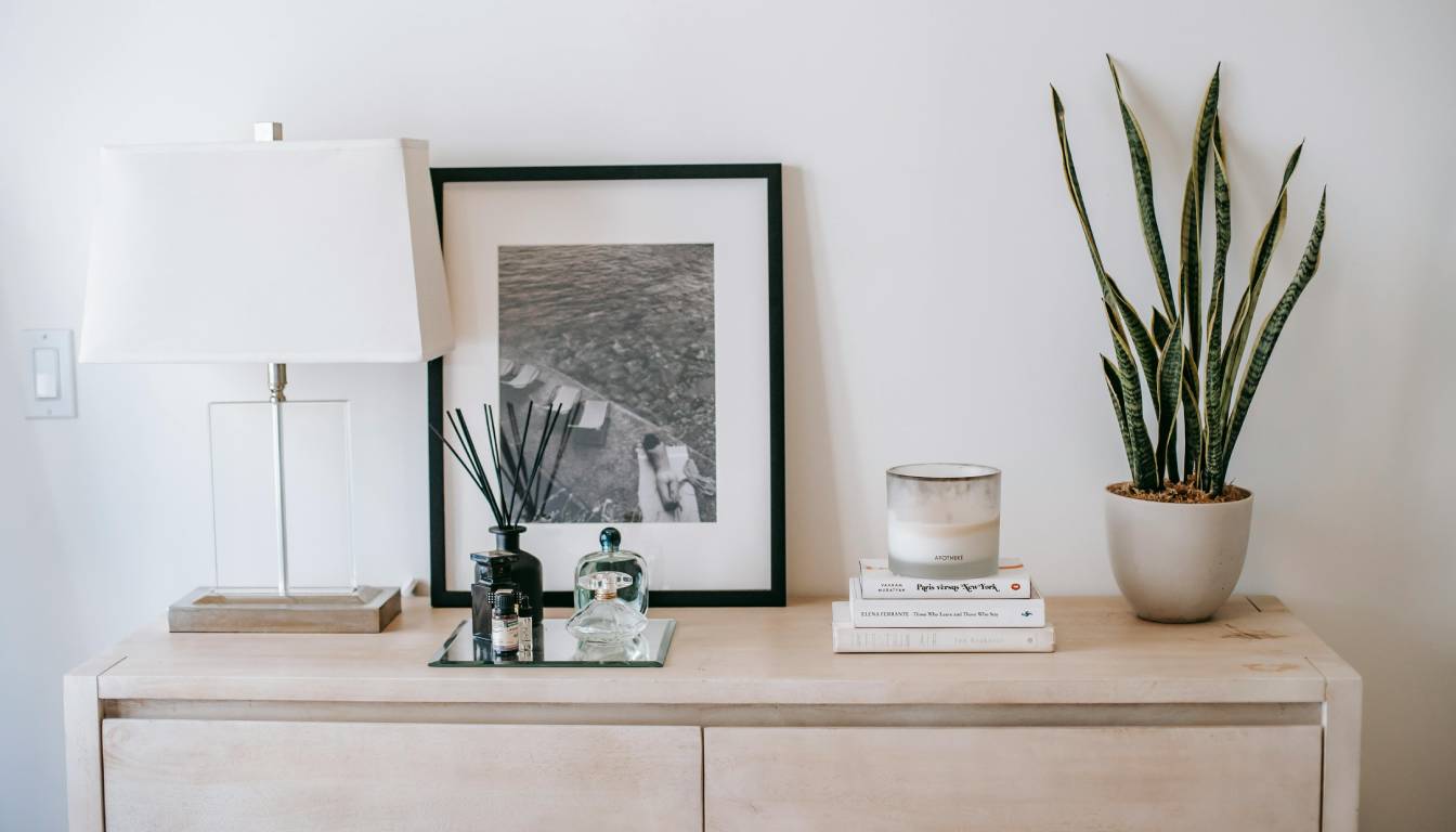 A modern wooden dresser with a variety of decorative items including a white lamp, a black and white framed photograph, a tray with small bottles, a stack of books, a candle, and a potted snake plant.