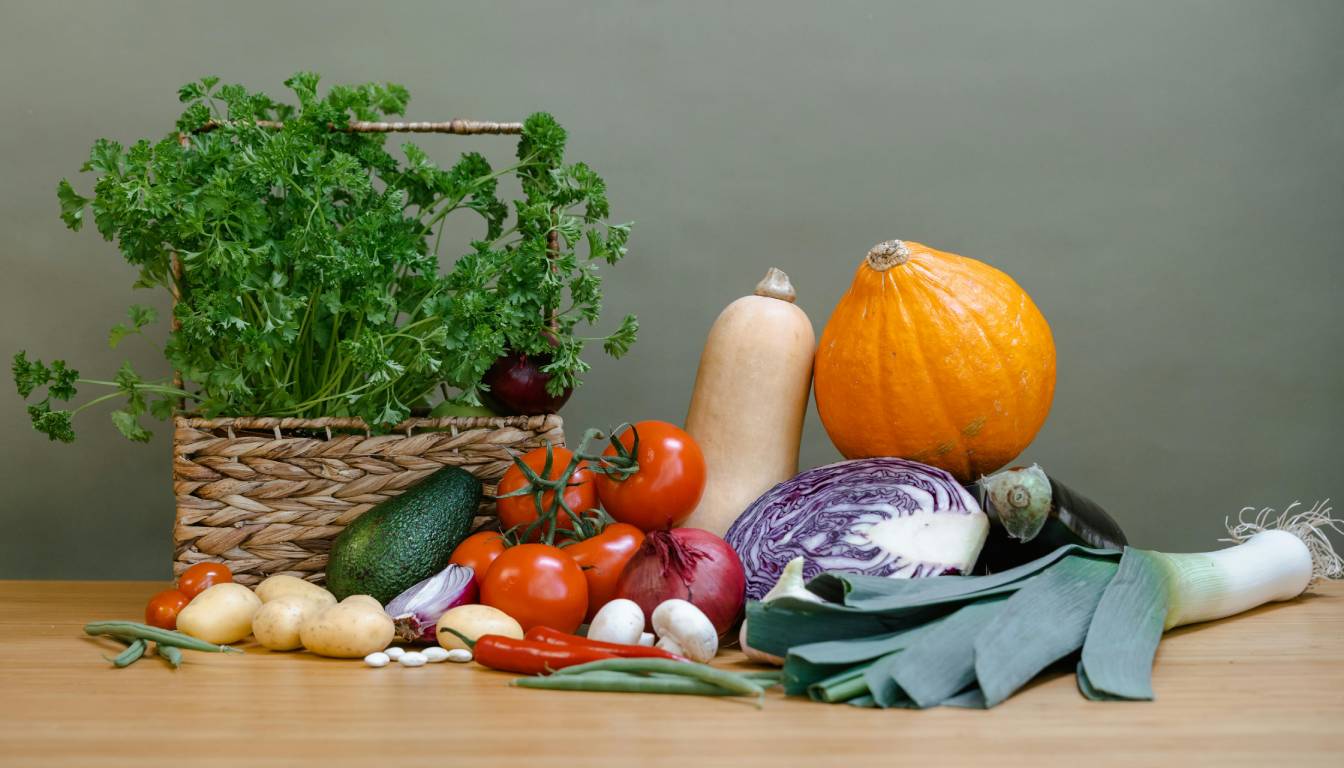 A variety of fresh vegetables and herbs displayed on a wooden surface, including parsley, tomatoes, squash, pumpkin, cabbage, leeks, potatoes, avocado, red onion, green beans, chili peppers, and garlic.