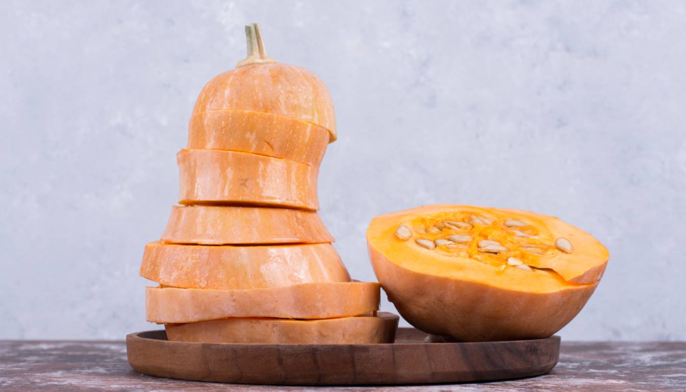 Sliced butternut squash stacked on a wooden plate next to a halved butternut squash showing seeds.