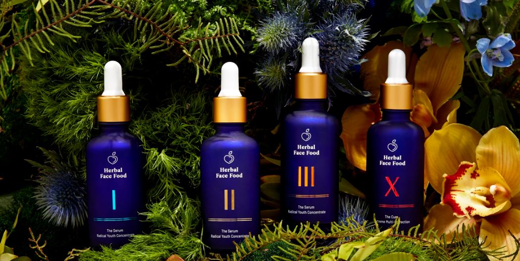 Four bottles of “Herbal Face Food” serums are displayed amidst lush greenery and vibrant flowers, highlighting the product’s natural ingredients.