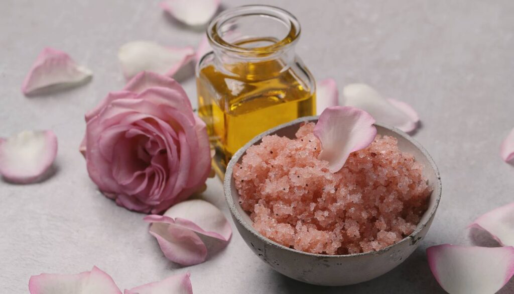 A bowl of pink bath salts with a pink rose petal on top, a glass bottle of yellow oil, and scattered pink rose petals on a light gray surface.