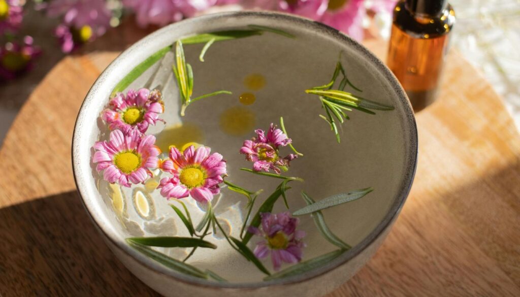A bowl of water with floating pink flowers and green herbs, with an amber glass bottle in the background.