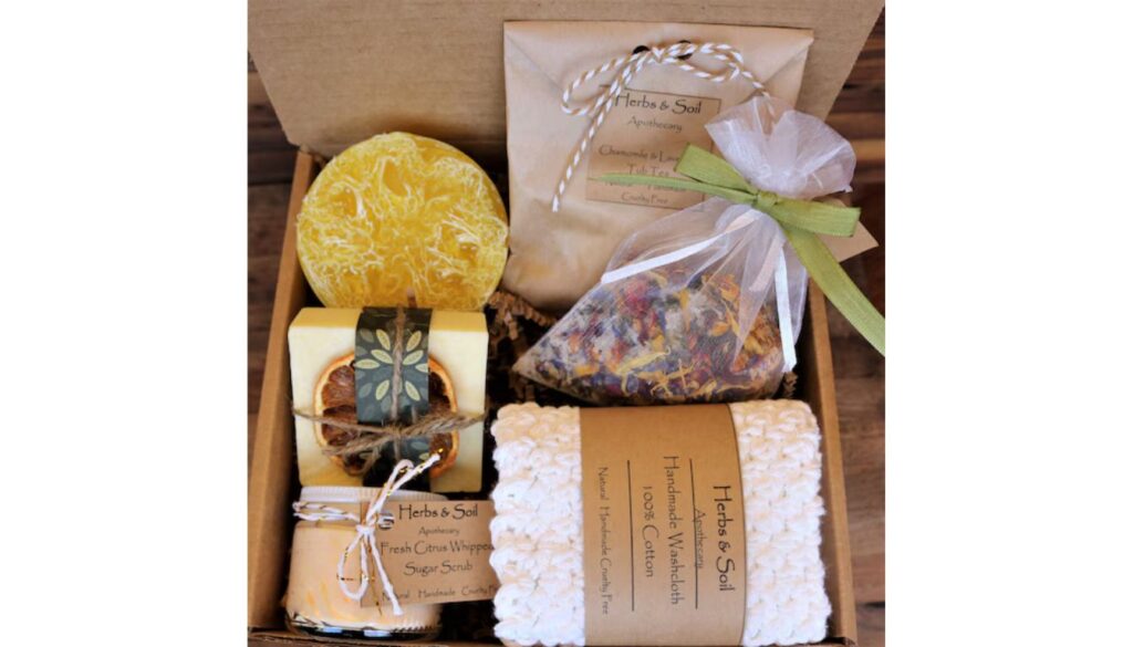 A beautifully arranged gift box containing herbal and natural products, including a loofah sponge, soap, dried flowers, sugar scrub, and a handmade washcloth.