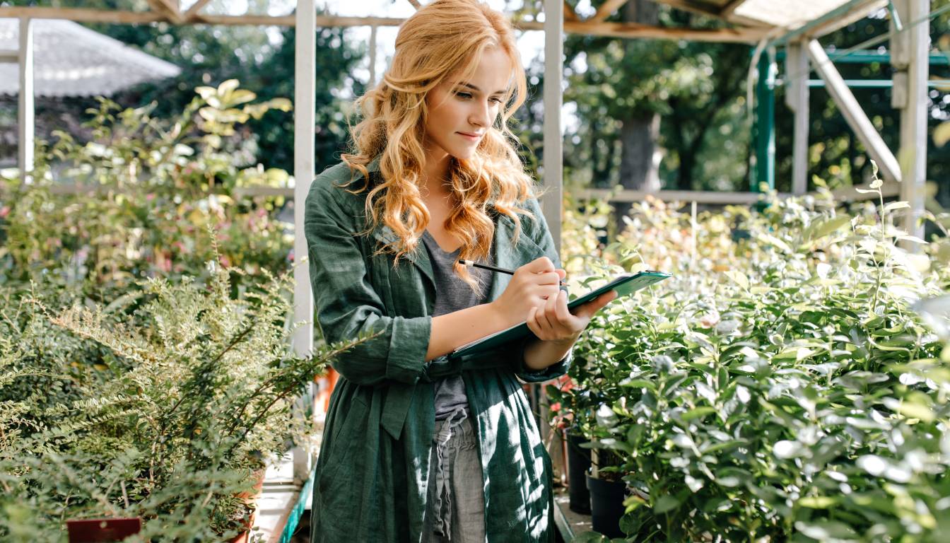 A person with long hair, wearing a green jacket, is taking notes on a clipboard while standing among various plants in a greenhouse.