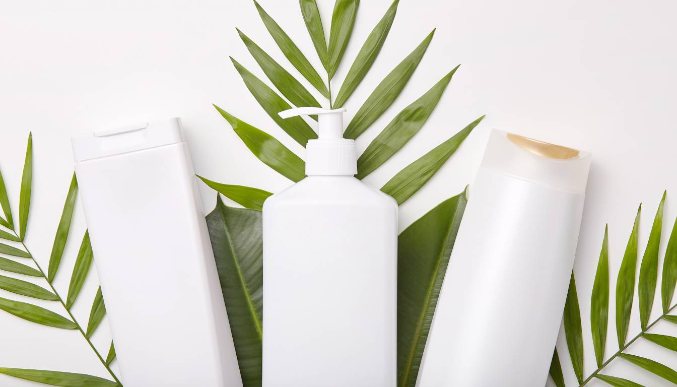 Three white shampoo and lotion bottles arranged on green palm leaves, representing natural hair care products.