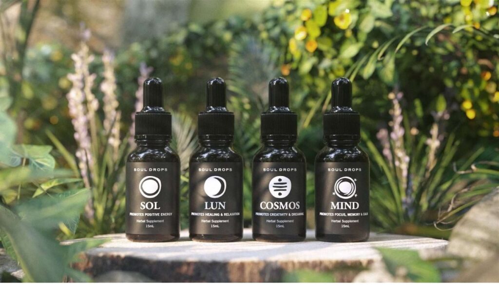 Four bottles of Soul Drops herbal supplements labeled SOL, LUN, COSMOS, and MIND, placed outdoors among greenery.