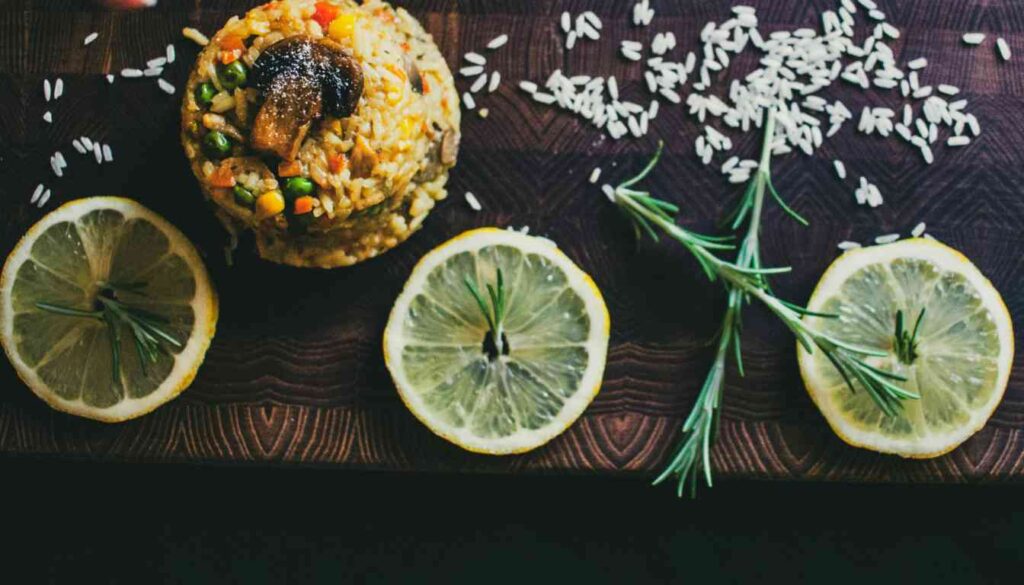 A vibrant dish of herb lemon rice garnished with fresh herbs and lemon slices on a dark wooden surface, surrounded by scattered rice grains and rosemary sprigs.