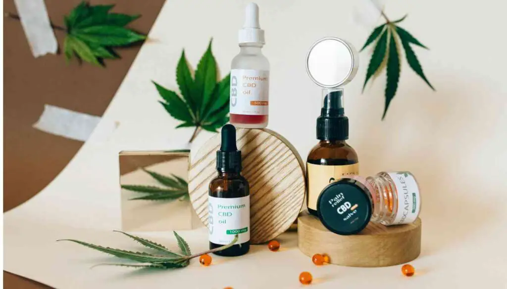 A display of cannabis tincture products with a backdrop of marijuana leaves. The image features a spray bottle, dropper bottle, and a jar labeled ‘Premium CBD’ on wooden stands, alongside scattered orange capsules and a magnifying glass.