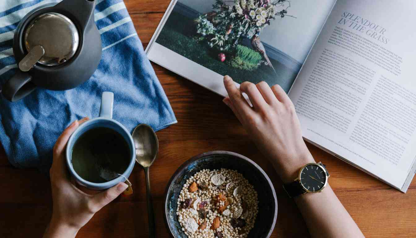 A person’s hands holding a cup of herbal tea over a wooden table, with an open magazine featuring an article on herbs, a bowl of granola, and a teapot.