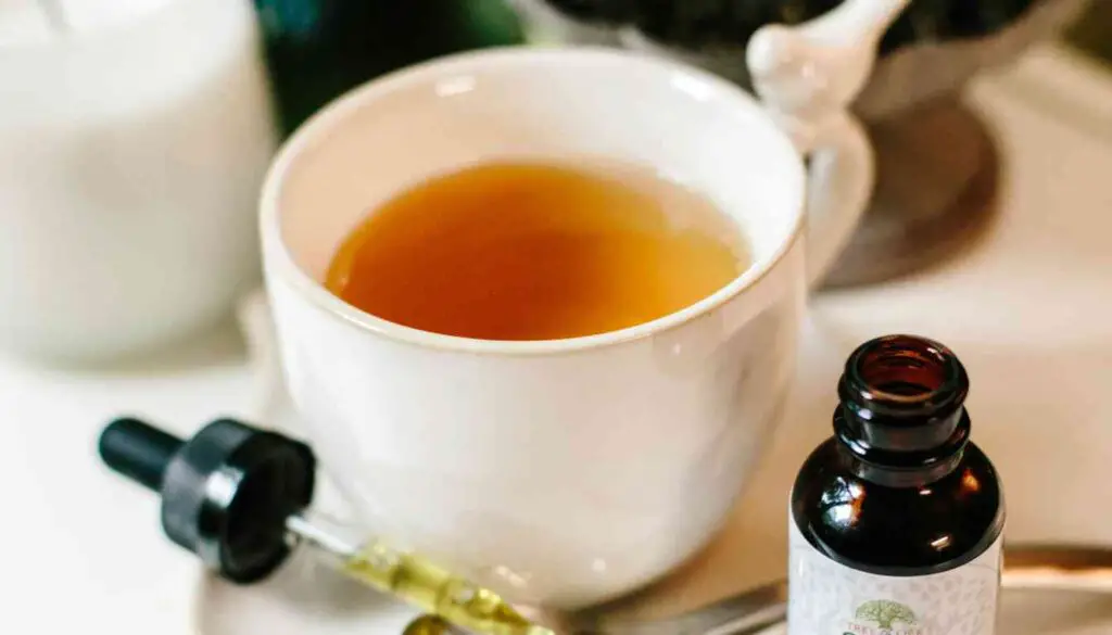 A small brown glass bottle with a dropper filled with a yellow liquid, labeled “Leanor Herbal Slimming Drops,” is placed next to a white ceramic cup filled with tea on a white surface. The bottle has a green leaf symbol on it.