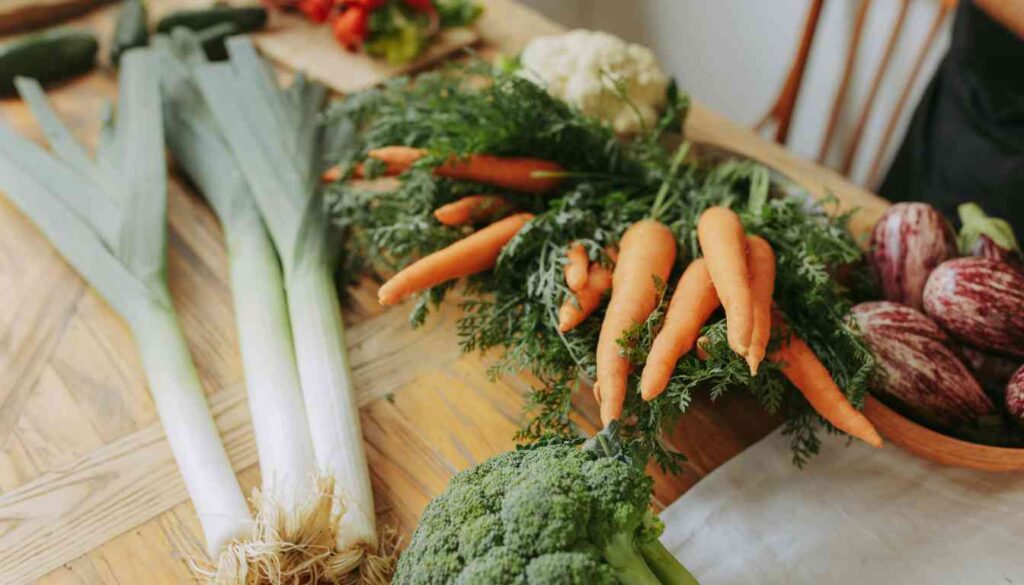 A variety of fresh vegetables including leeks, carrots, broccoli, cauliflower, and eggplants are arranged on a wooden table.