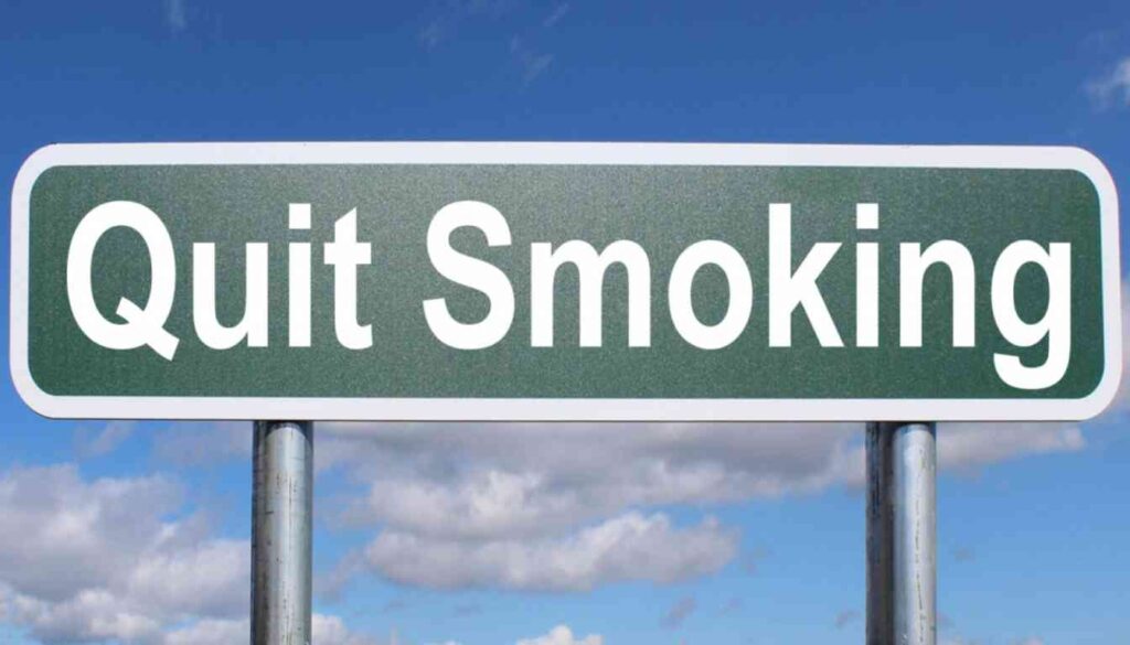 A road sign with the words “Quit Smoking” against a backdrop of a clear blue sky with scattered clouds.