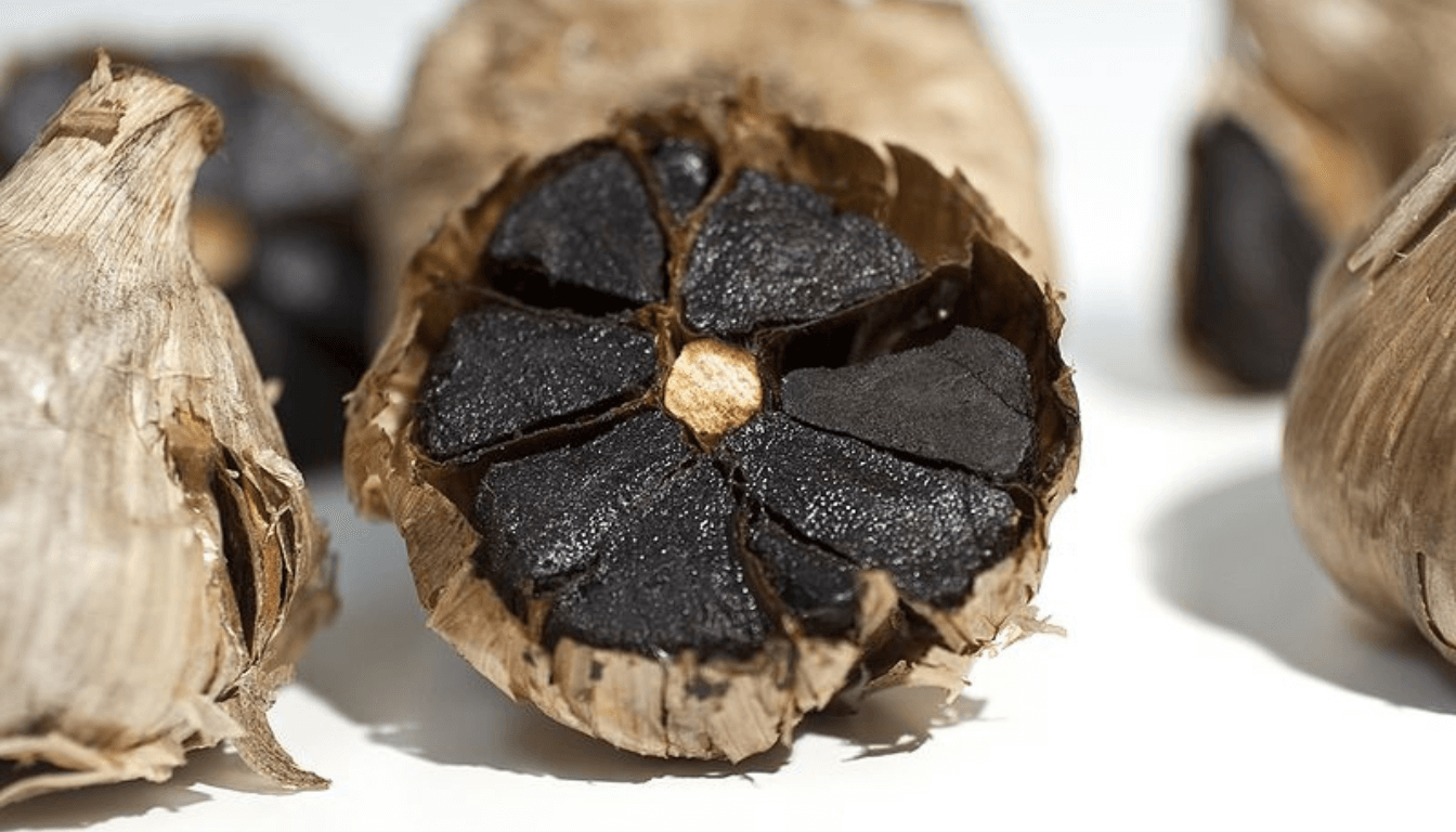 A close-up image of a bulb of black garlic with its cloves exposed, alongside two intact bulbs, against a white background.