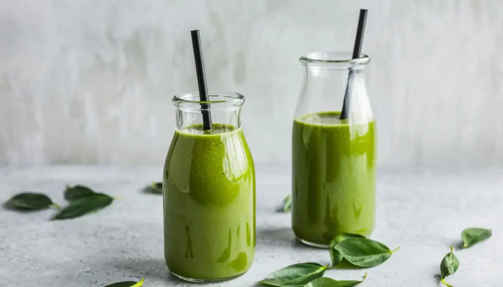 Two bottles of vibrant green herbal juice with black straws, surrounded by fresh green leaves on a light grey surface.