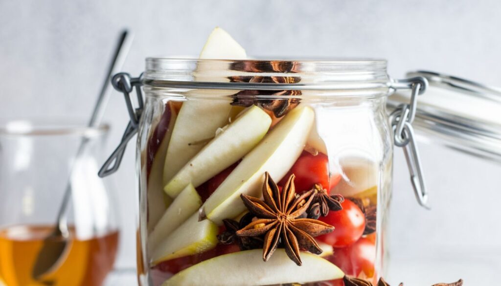 A clear glass jar with a metal clasp lid, filled with various ingredients including apple slices, whole star anise, and red chili peppers.