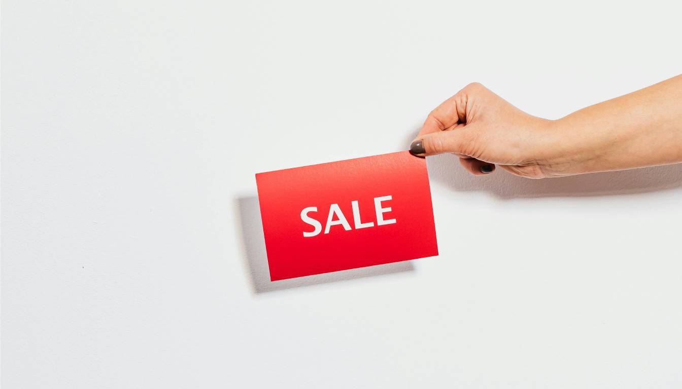 A hand holding a red card with the word "SALE" written in white, against a plain white background.