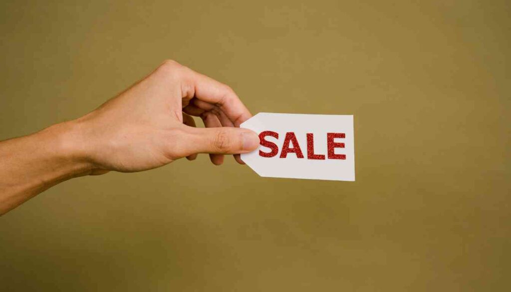 A hand holding a white card with the word “SALE” printed in red capital letters, against a solid beige background.