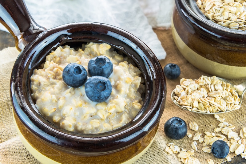 A bowl of creamy oatmeal topped with three fresh blueberries, with a spoonful of raw oats and additional blueberries scattered around.