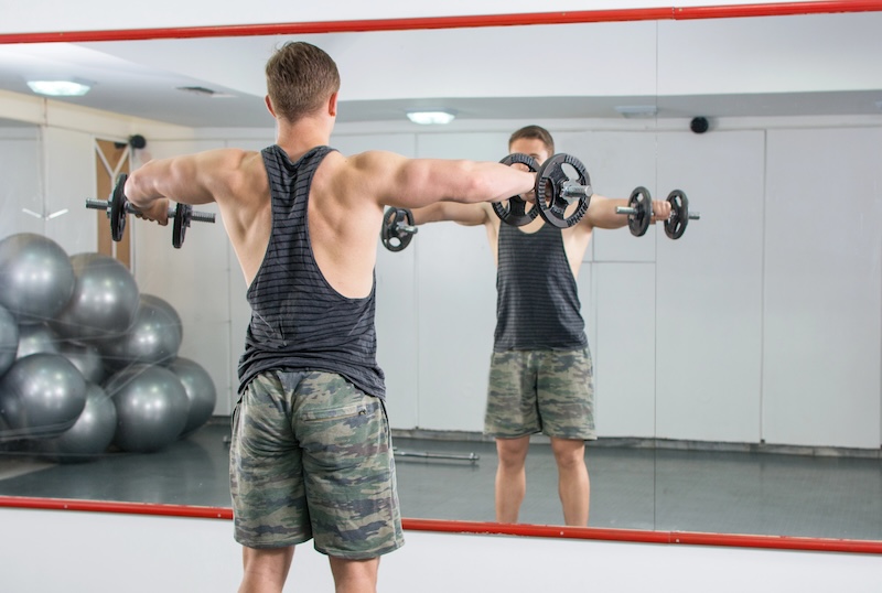 A person in a gym performing lateral raises with dumbbells in front of a mirror.