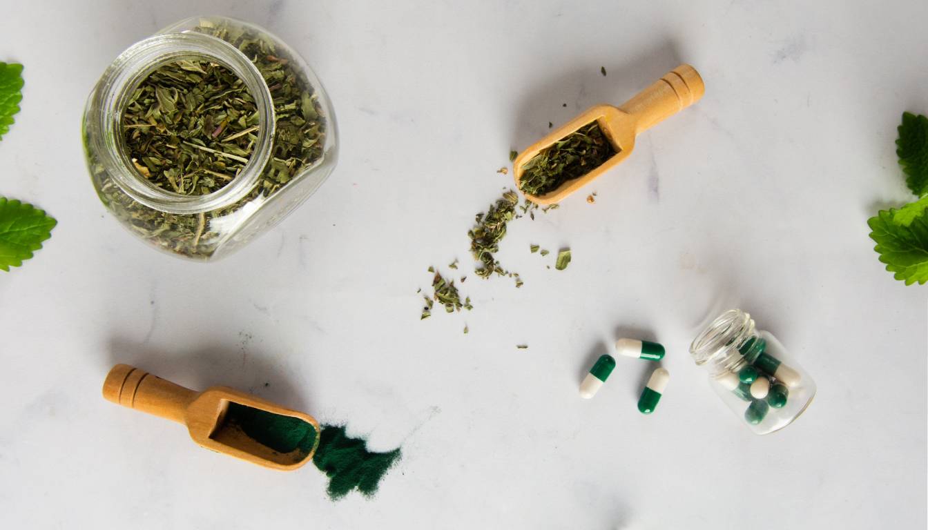 A flat lay image showing a jar of dried herbs, two wooden scoops with herbs and green powder, and a small glass jar with green and white capsules, placed on a white surface with green leaves around.