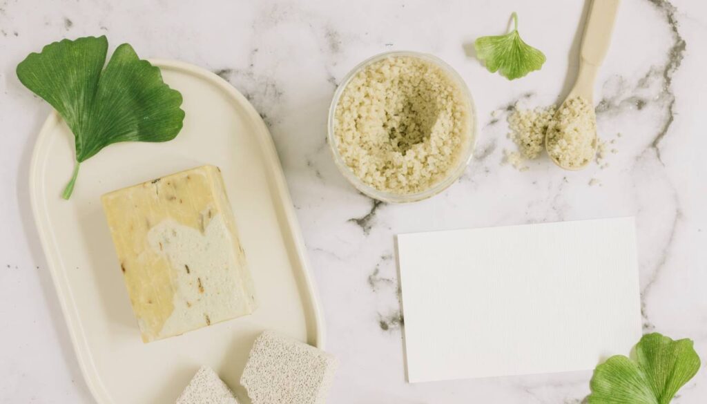 A bar of cilantro-scented soap on a tray, accompanied by a jar of exfoliating scrub, a spoon, and green leaves, all arranged on a marble surface.