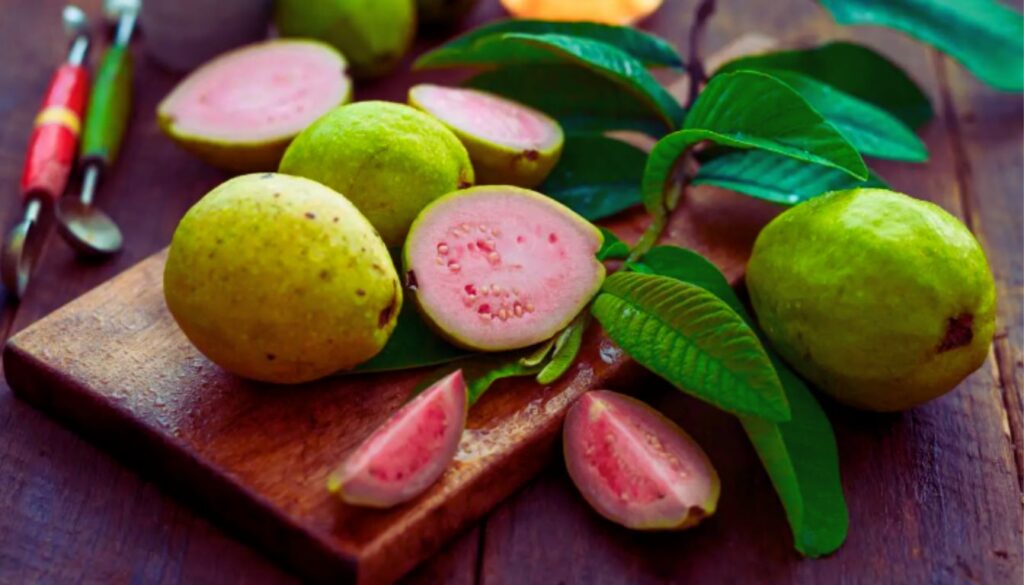 Fresh guavas, some whole and some cut open, displayed on a wooden cutting board with green leaves and kitchen utensils in the background.