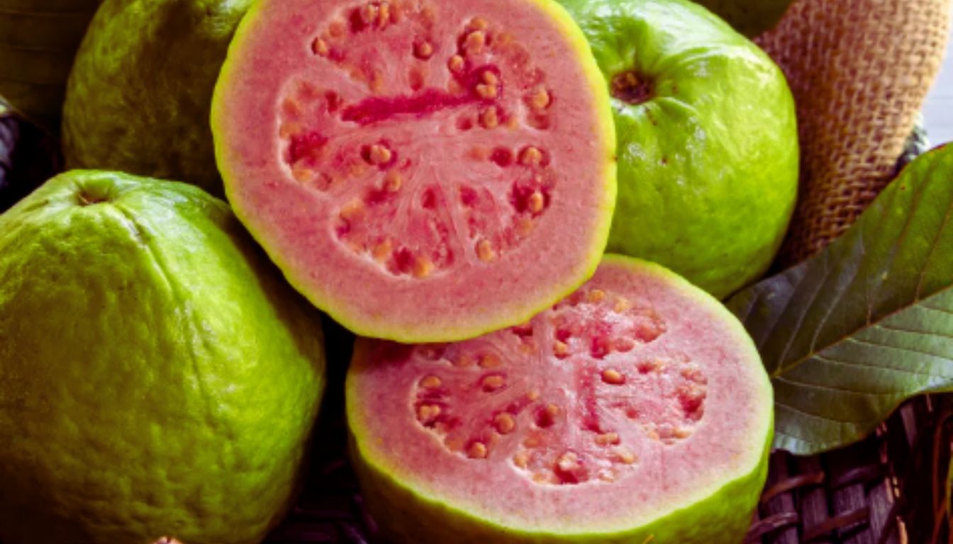 A close-up of several guavas, with one guava cut in half to reveal its pink flesh and numerous seeds.