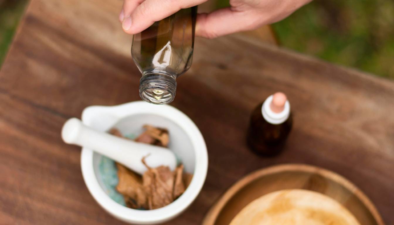 A person pouring liquid from a brown bottle into a mortar containing dried herbs, with a pestle resting inside the mortar.