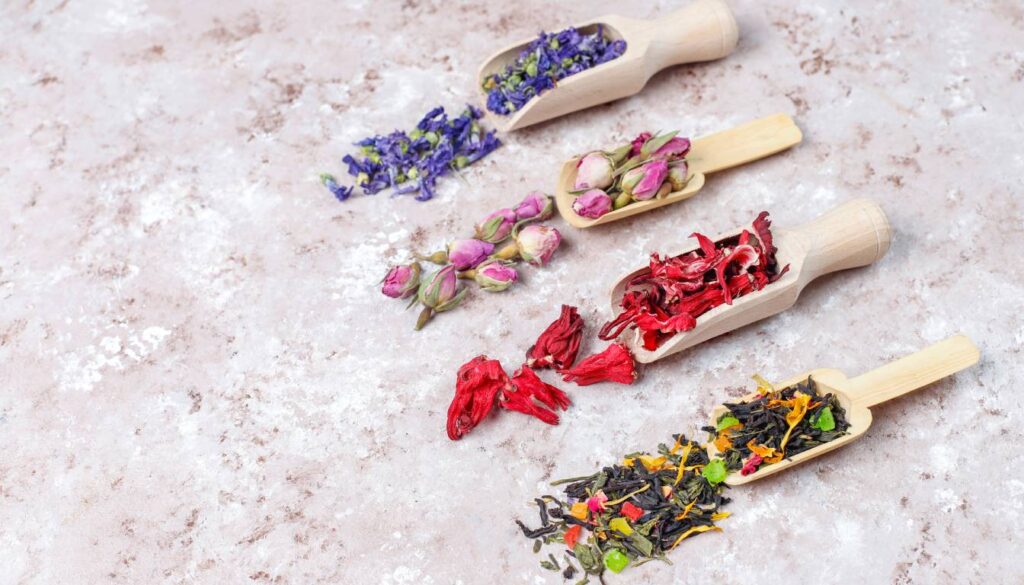Various dried herbs and flowers in wooden scoops on a textured surface.